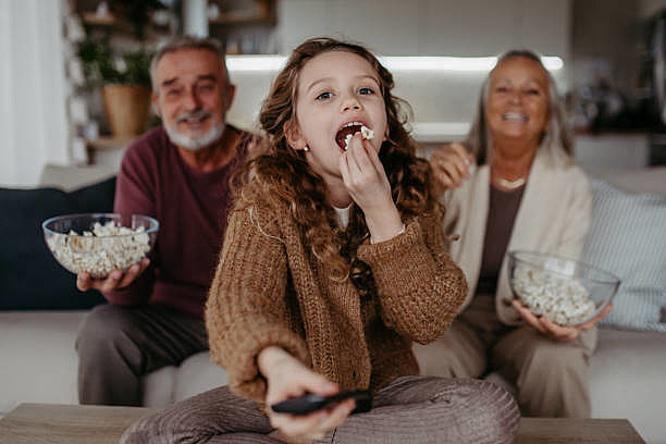 Grandparents watching TV with their granddaughter and eating popcorn.